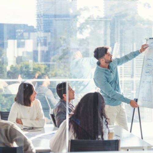 Business people watching a presentation on the whiteboard. A man is writing on the whiteboard with charts and graphs. They are sitting in a board room, there are laptop computers and technology on the table. All are casually dressed. There is a window behind him with city views.